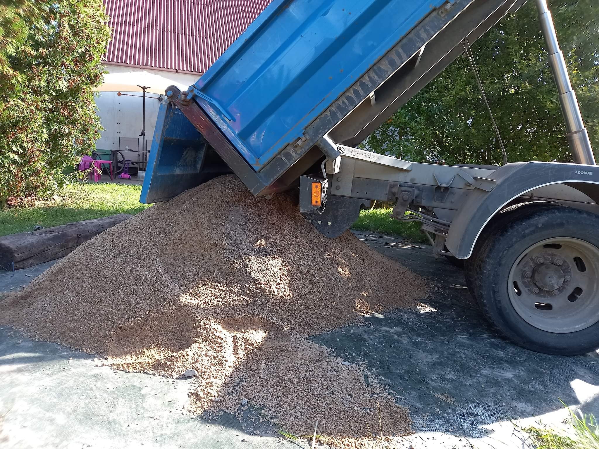 Wyładowany z wywrotki piasek na czarnej macie, widoczny fragment ogrodu z domem w tle.