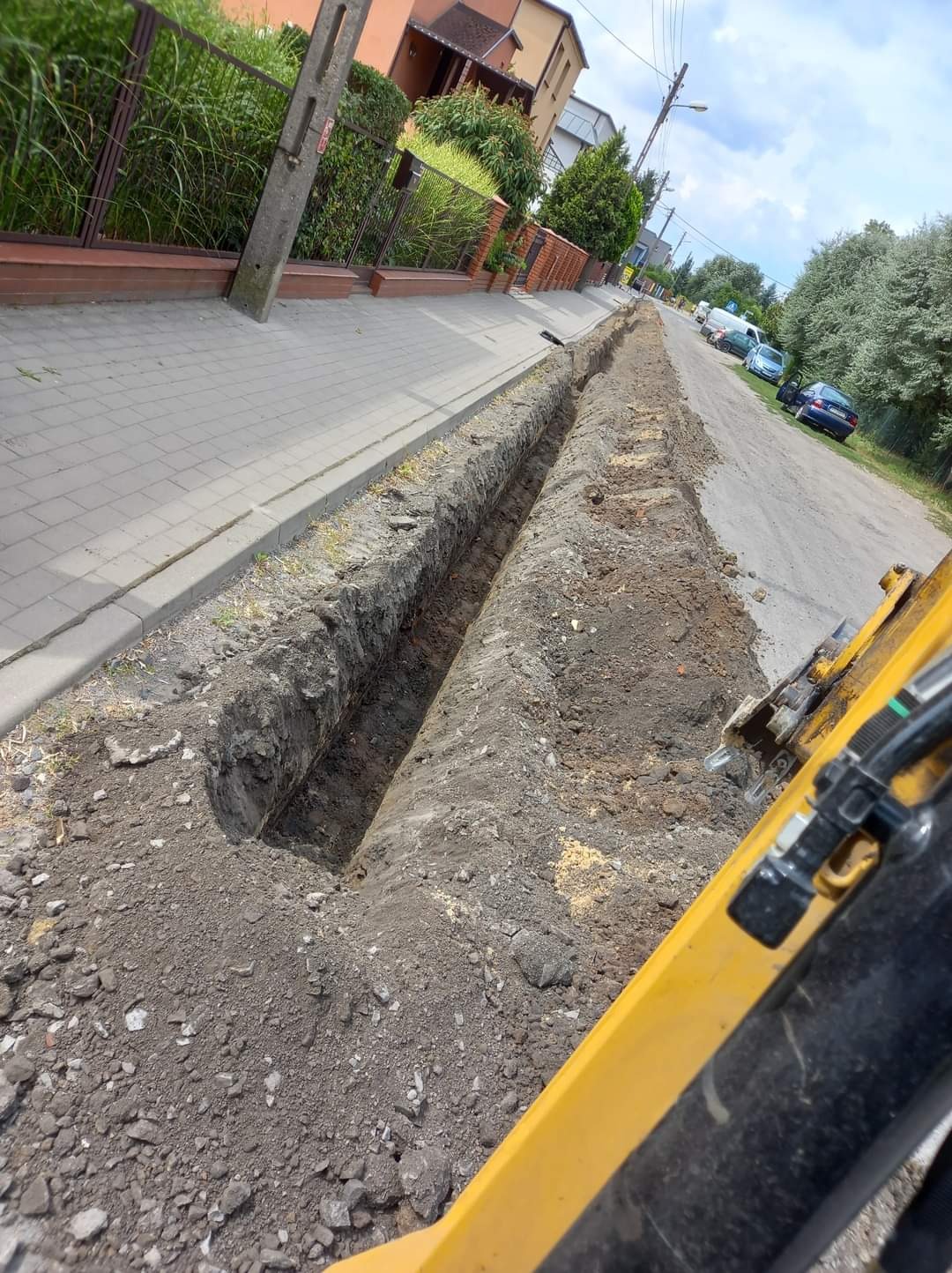 Długi, wąski wykop wzdłuż chodnika i ulicy, wykonany koparką widoczną w prawym dolnym rogu, z odsłoniętą ziemią i kamieniami.