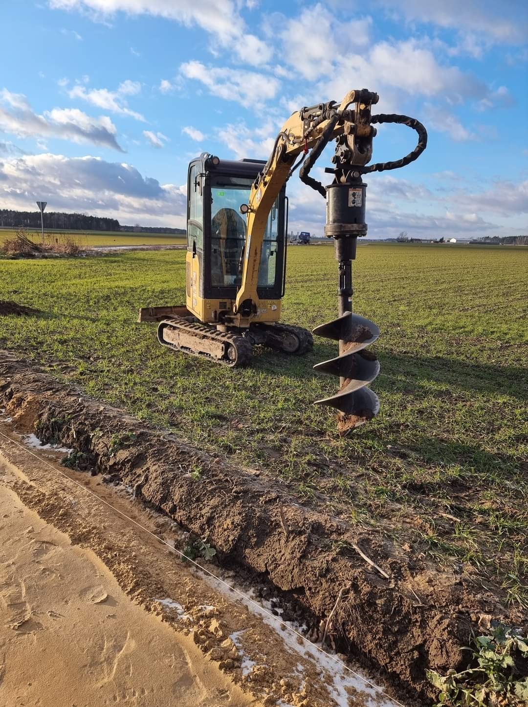 Żółta mini koparka z wiertłem ziemnym w trakcie pracy na polu, obok wykopany rów z widoczną linką wyznaczającą linię, błękitne niebo z chmurami w tle.