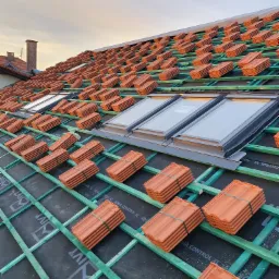 Układanie czerwonej dachówki karpiówki na nowym dachu z oknami połaciowymi, widoczna impregnacja i zielona konstrukcja więźby dachowej.