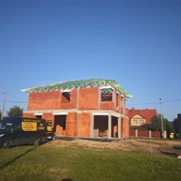 Budowa domu jednorodzinnego w trakcie wznoszenia więźby dachowej, z widocznym samochodem dostawczym firmy budowlanej z reklamą na boku. Czerwona cegła porotherm, zielona impregnacja drewna więźby.