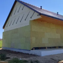 Budynek w trakcie ocieplania, widoczne płyty izolacyjne na ścianach i membrana na dachu z ciemną dachówką, w tle błękitne niebo.