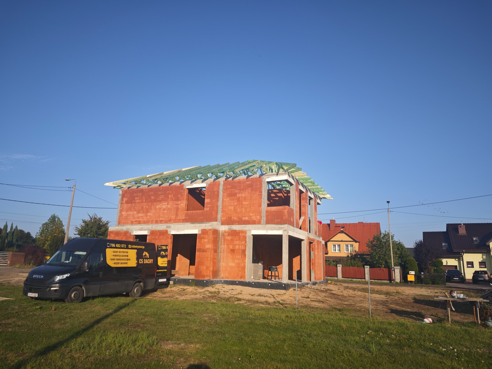 Budowa domu jednorodzinnego w trakcie wznoszenia więźby dachowej, z widocznym samochodem dostawczym firmy budowlanej z reklamą na boku. Czerwona cegła porotherm, zielona impregnacja drewna więźby.