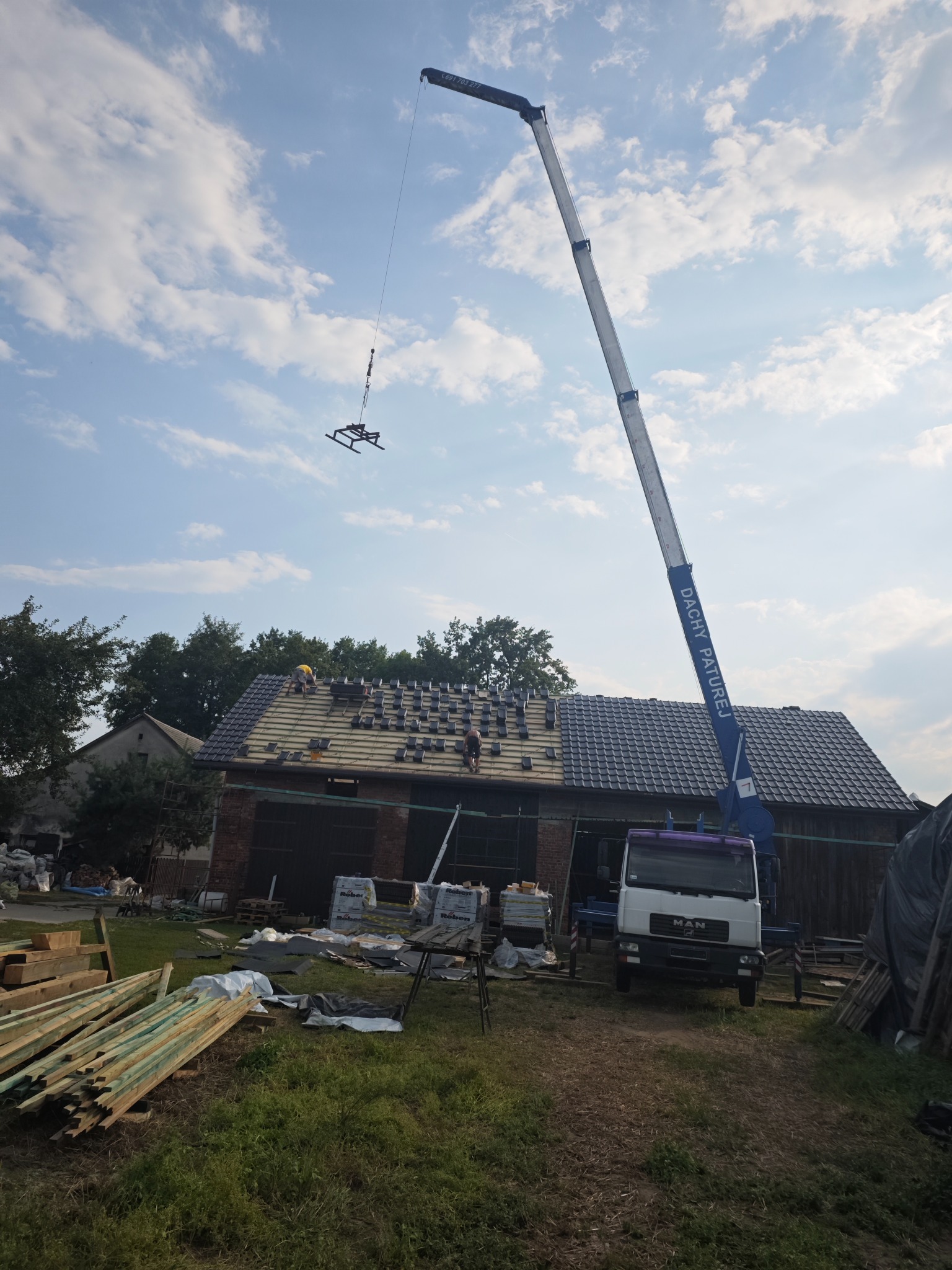 Montaż pokrycia na dachu budynku gospodarczego przy użyciu dźwigu MAN z wysięgnikiem, widoczne ułożone dachówki i pracownicy w trakcie robót, na ziemi przygotowane drewno i materiały budowlane.