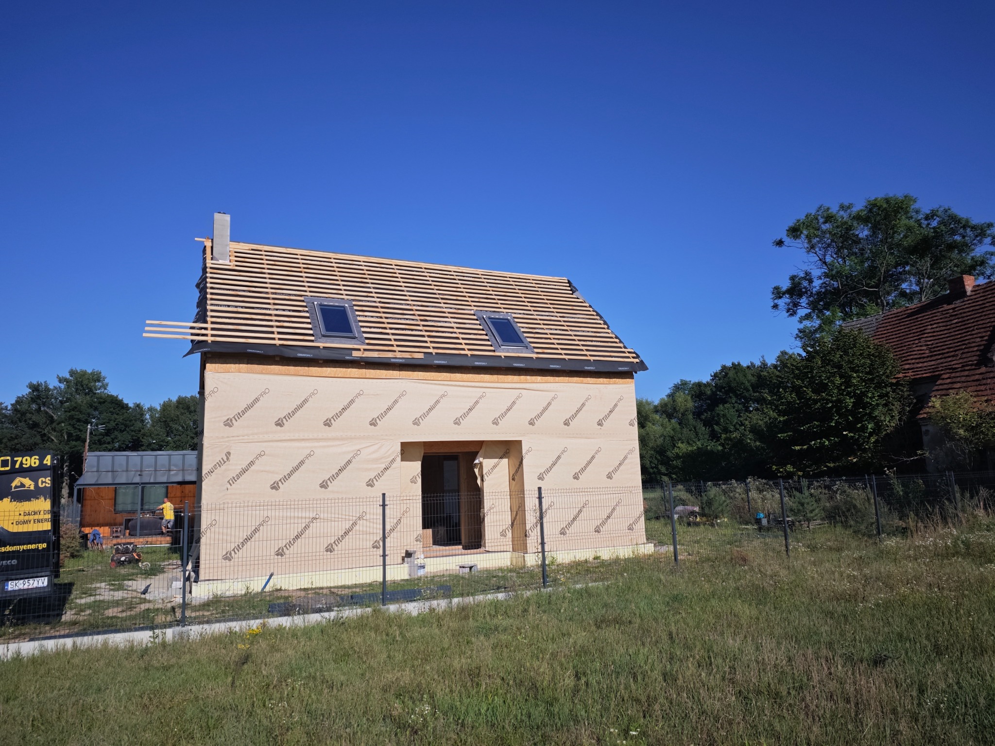 Budynek w trakcie budowy z widoczną więźbą dachową, deskowaniem i oknami dachowymi, zabezpieczony membraną TitaniumPRO, na tle błękitnego nieba.