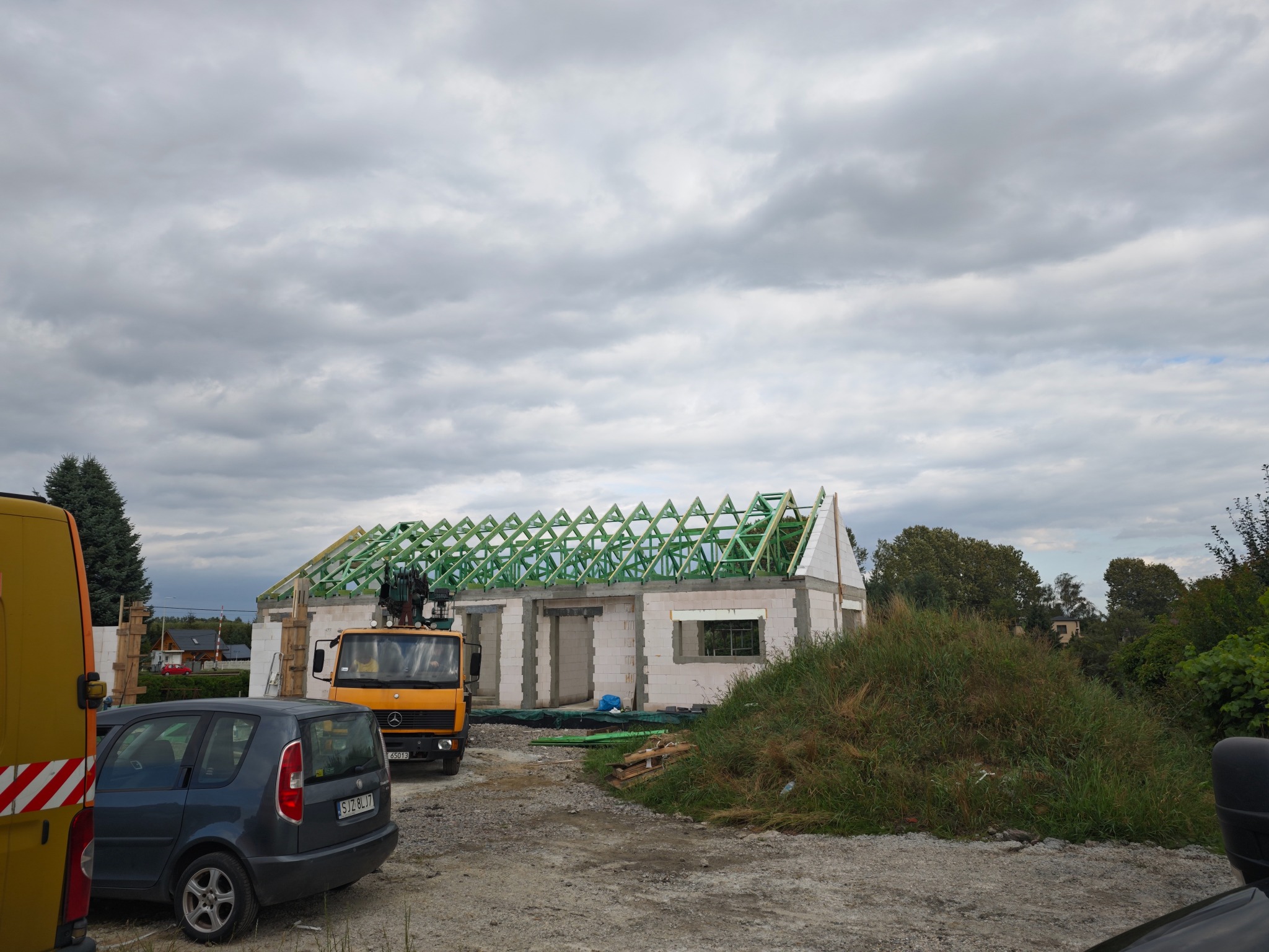Budowa domu jednorodzinnego w trakcie wznoszenia konstrukcji nośnej dachu; widoczna więźba dachowa, ściany z pustaków oraz auto z dźwigiem i samochody osobowe na placu budowy, pochmurne niebo w tle.