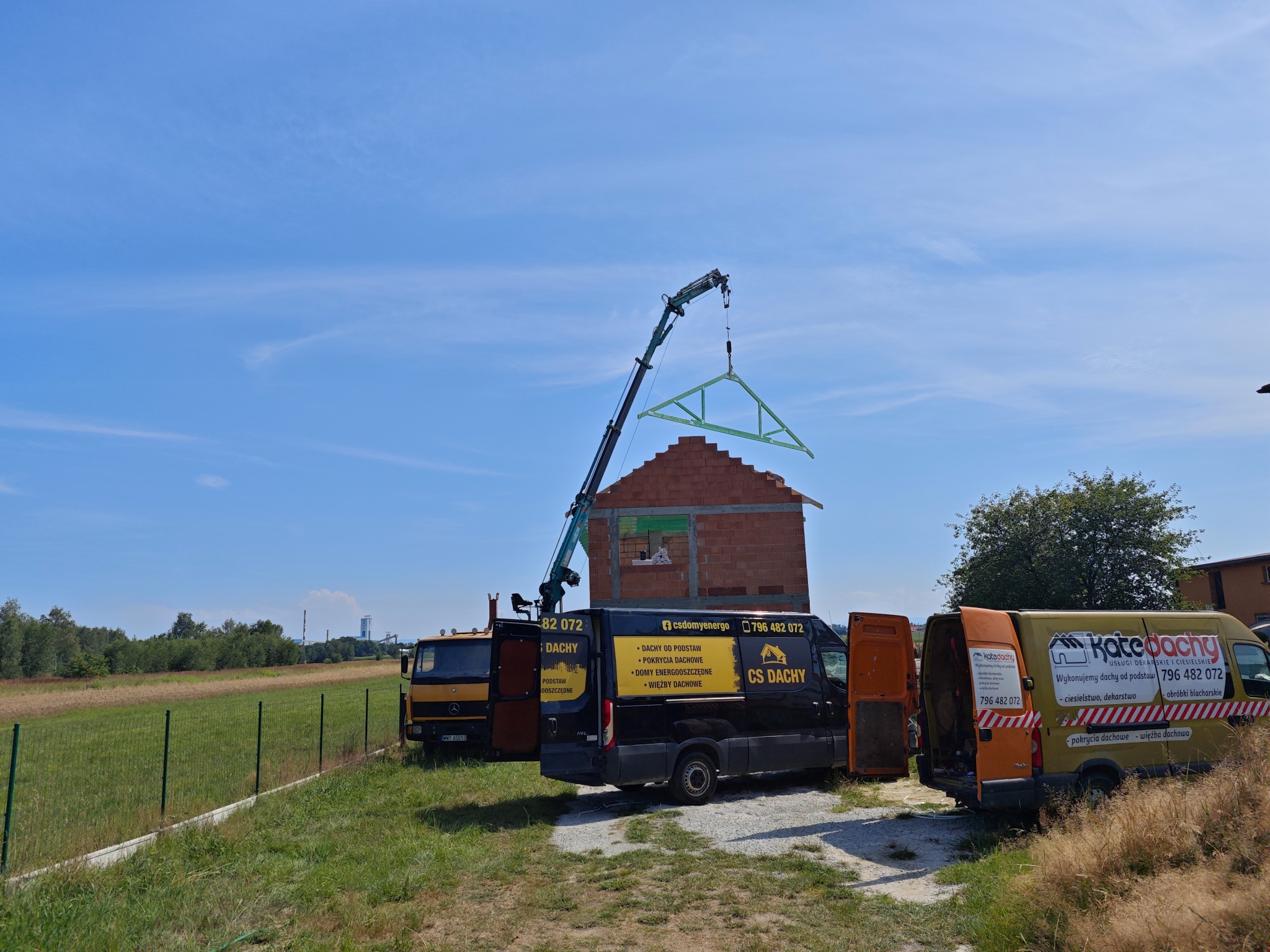 Montaż więźby dachowej na budowie domu jednorodzinnego przy użyciu dźwigu, w tle pola i lasy, na pierwszym planie samochody firm dekarskich z reklamami na karoserii.