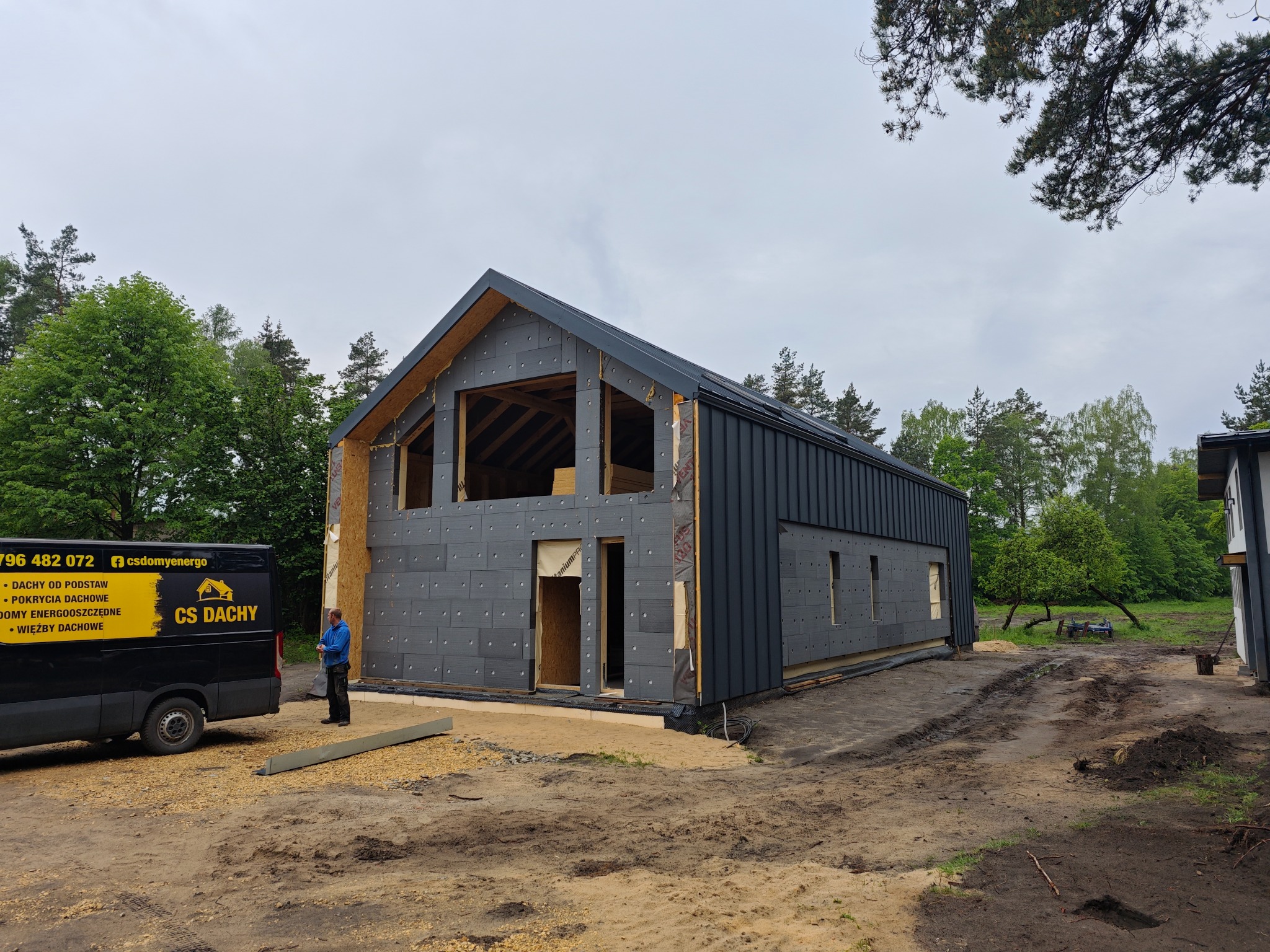 Budowa domu jednorodzinnego w trakcie montażu izolacji i blachy na dachu, widoczny pracownik firmy CS Dachy oraz samochód firmowy z numerem telefonu i logo na tle lasu.