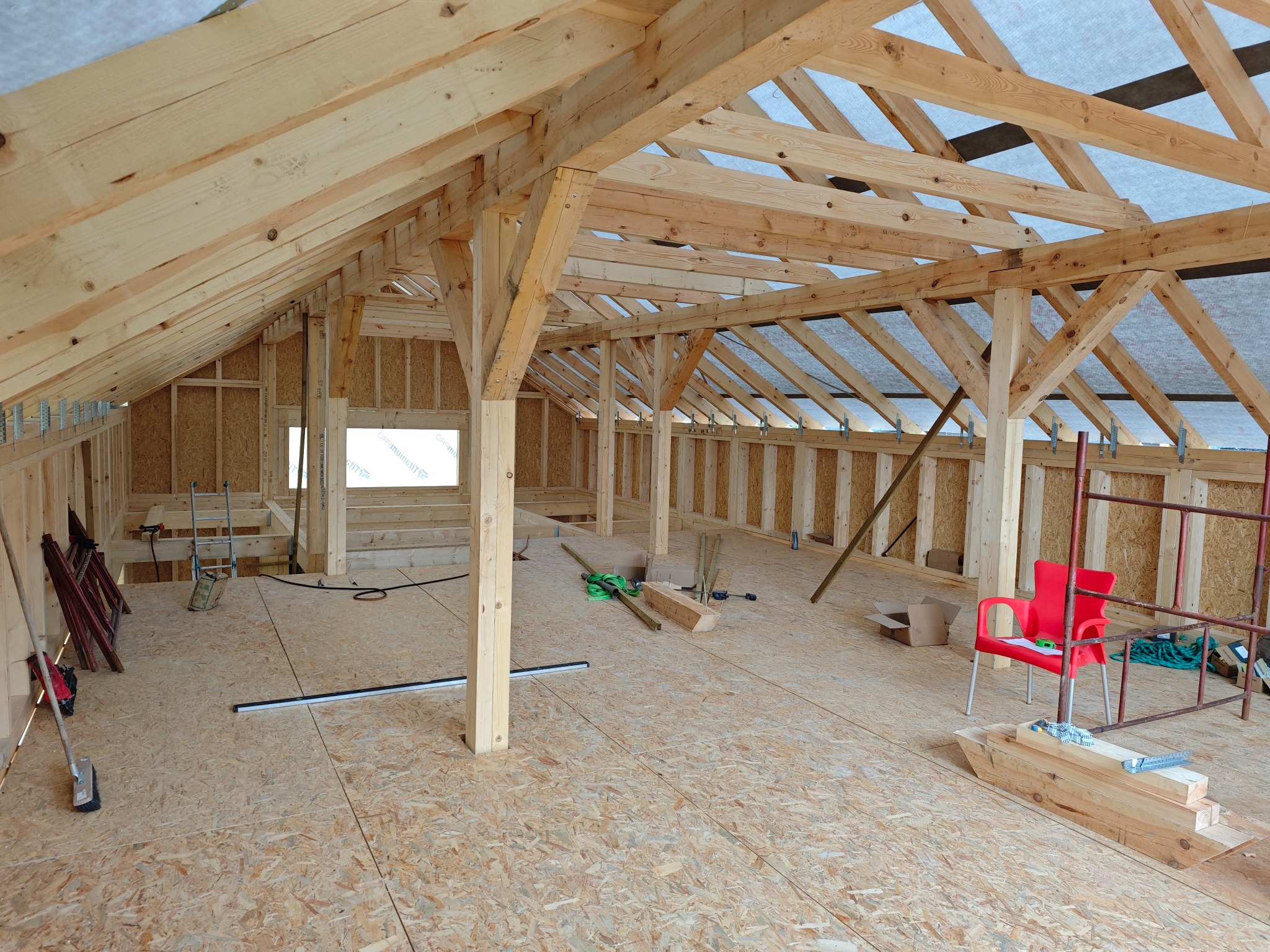 Wnętrze poddasza z widoczną drewnianą konstrukcją dachu, ścian i podłogi z płyt OSB, czerwone krzesło i rusztowanie w tle, narzędzia i materiały budowlane rozłożone na podłodze.