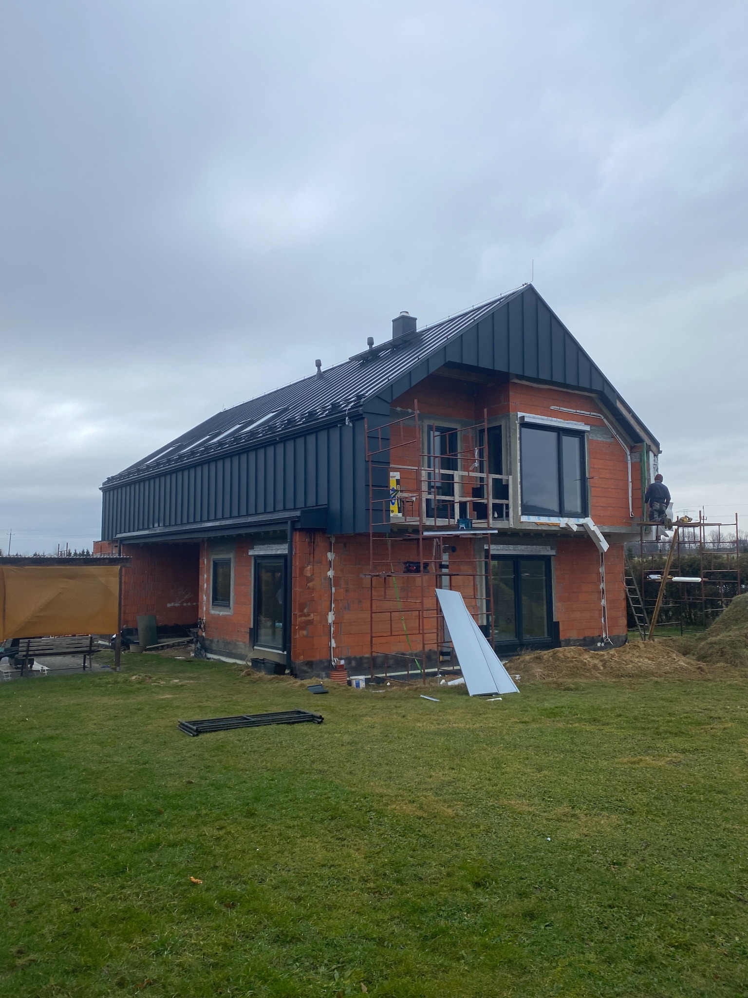 Nowoczesny dom w trakcie budowy z czarnym pokryciem, widoczne rusztowania, pracownik na rusztowaniu i elementy konstrukcyjne na trawniku.