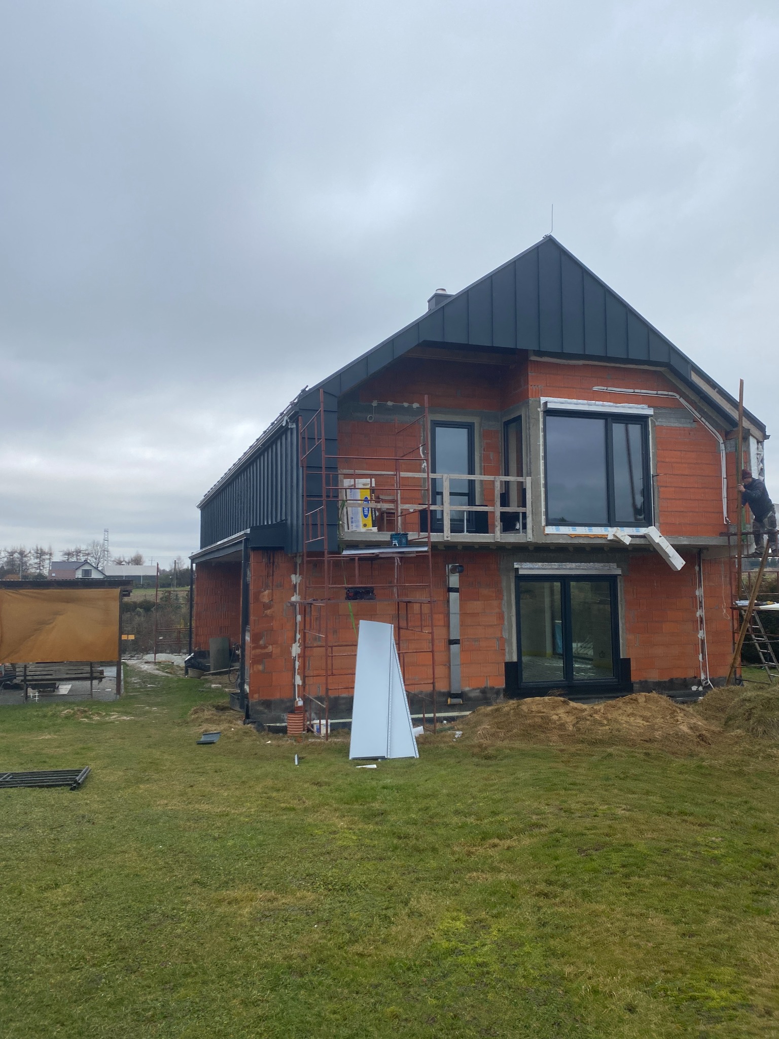 Budynek w trakcie budowy z zamontowaną blachą na dachu i pracownikiem na drabinie, widoczne rusztowania i ceglana konstrukcja.