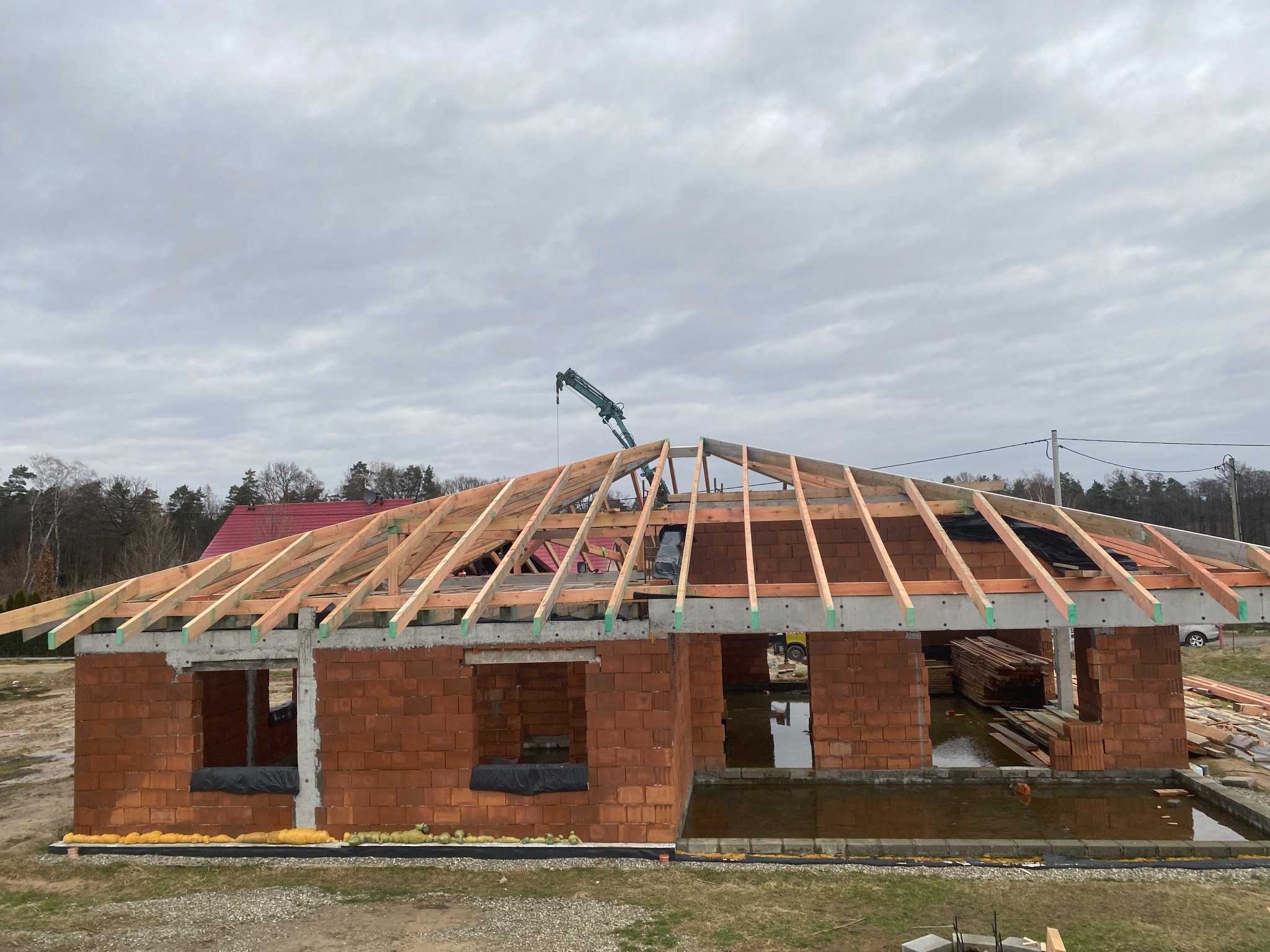 Budowa domu jednorodzinnego w stanie surowym otwartym: widoczna murowana konstrukcja z czerwonej cegły, betonowe nadproża i drewniana konstrukcja dachu z elementami podtrzymywanymi przez dźwig.