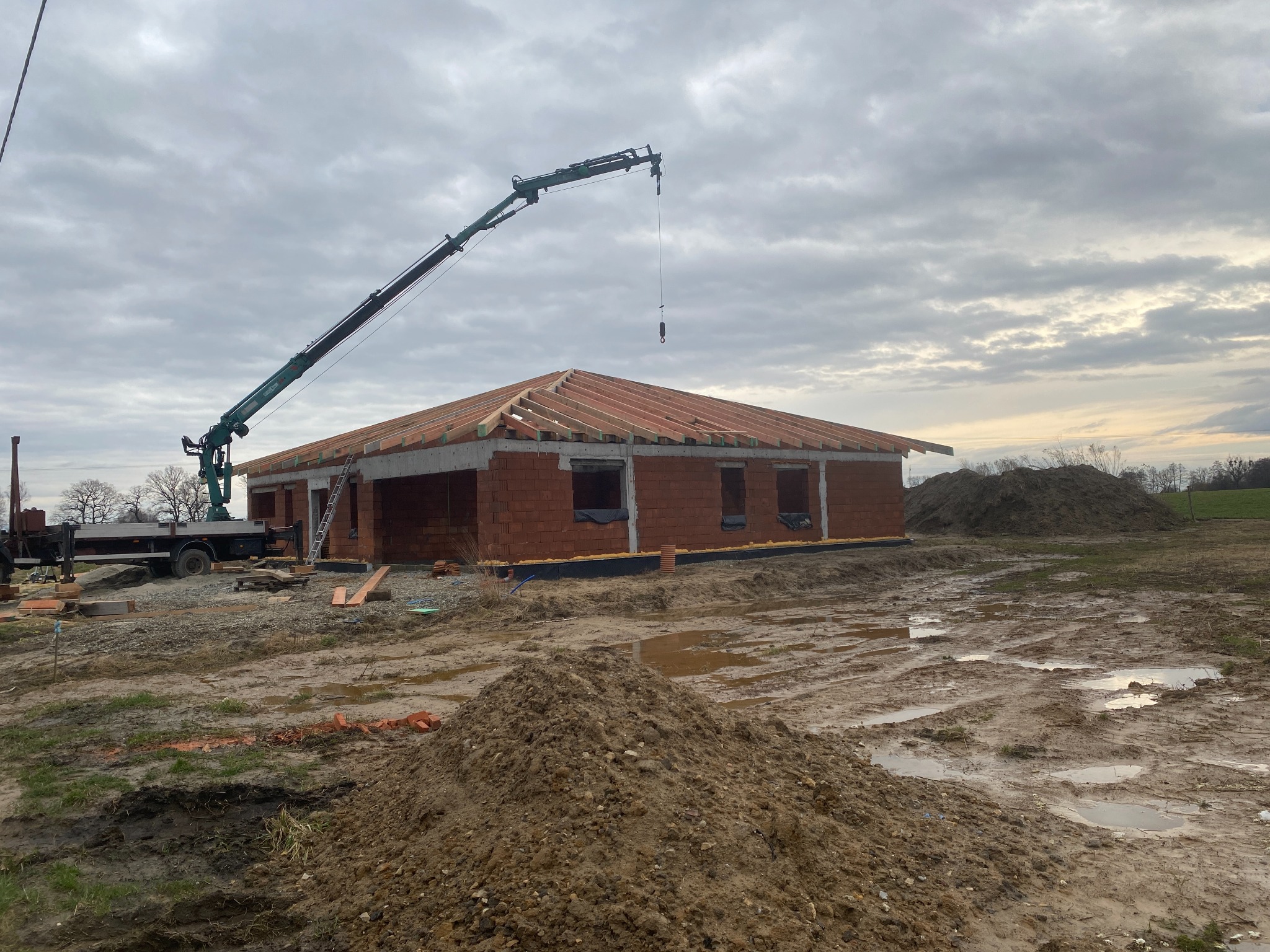 Budowa domu jednorodzinnego w trakcie: wzniesione mury z czerwonej cegły, drewniana konstrukcja przyszłego dachu, na placu budowy stoi dźwig, teren wokół rozkopany i błotnisty, pochmurne niebo.