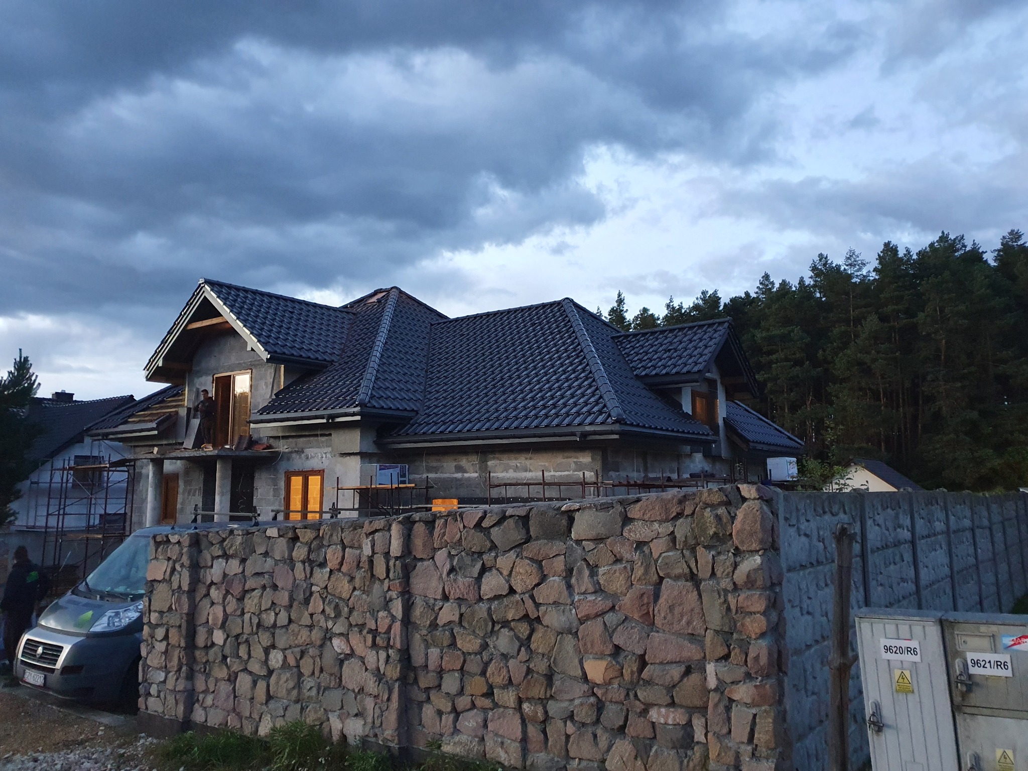 Dom w trakcie budowy z ciemną dachówką na dachu, widoczny pracownik na rusztowaniu, kamienny mur w pierwszym planie i pochmurne niebo w tle.