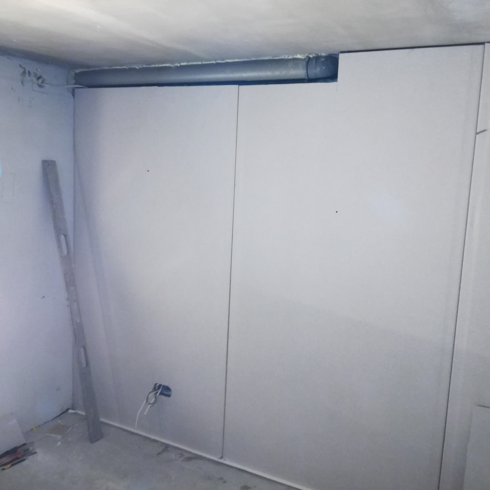 Wnętrze w trakcie remontu: ściana wyłożona płytami gipsowo-kartonowymi, widoczny poziomica oparta o ścianę, instalacja elektryczna wystająca ze ściany, rura wentylacyjna pod sufitem.