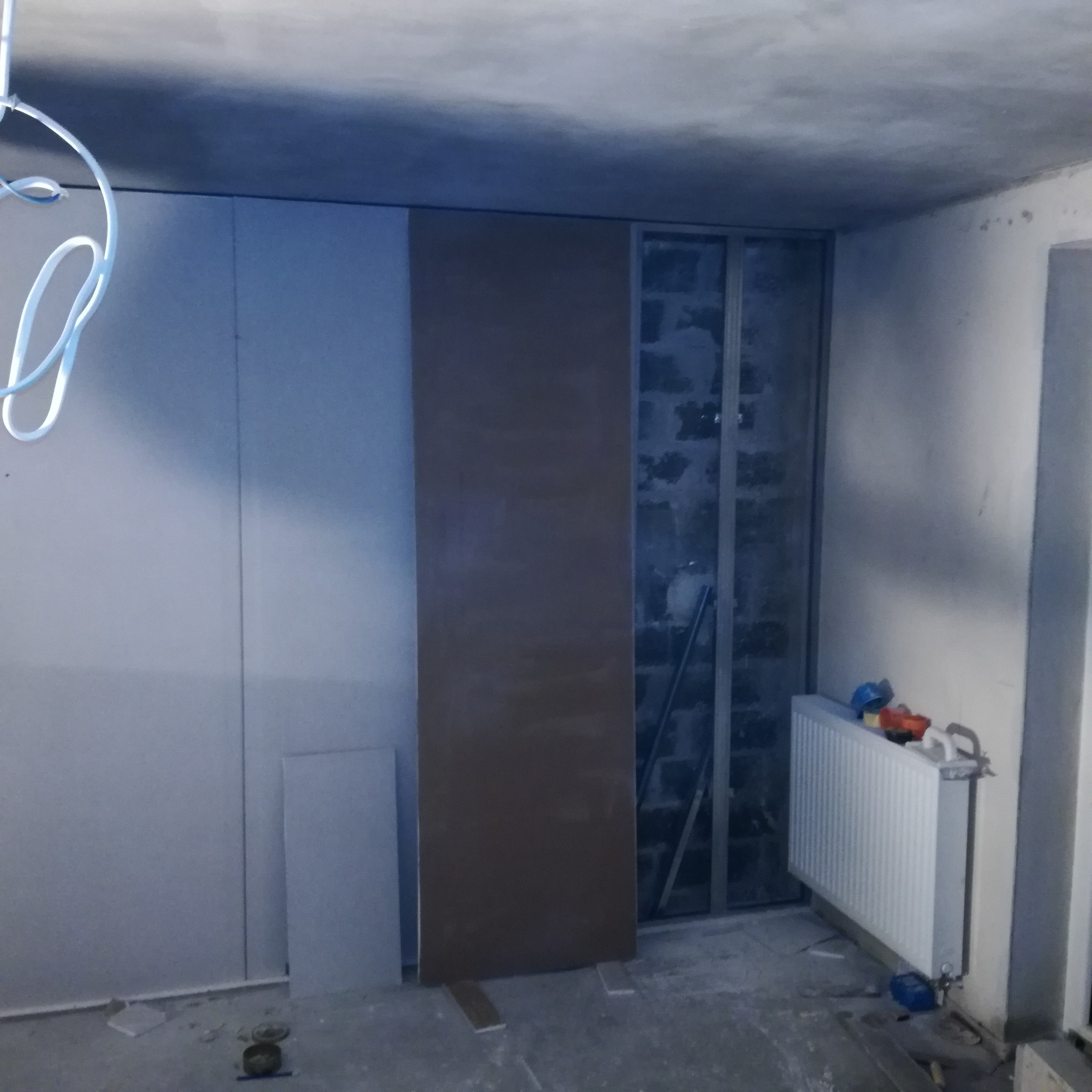 Wnętrze w trakcie remontu: ściany wyłożone płytami gipsowo-kartonowymi, widoczna konstrukcja metalowa, grzejnik i narzędzia na podłodze.