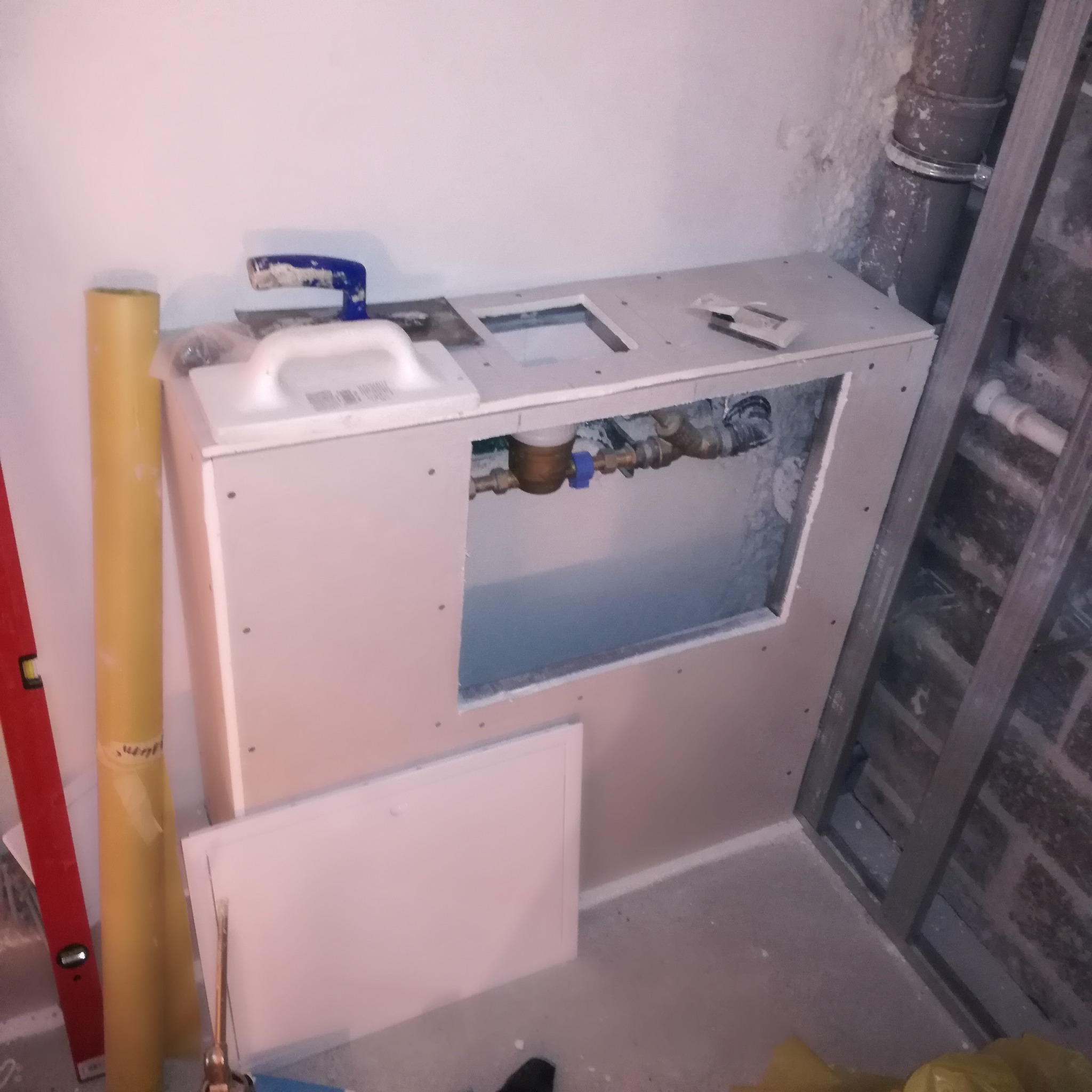 Montaż obudowy z płyty karton-gips nad instalacją wodną. Widoczne narzędzia: poziomica, szpachelki, wałek malarski. Otwór rewizyjny z dostępem do zaworów.