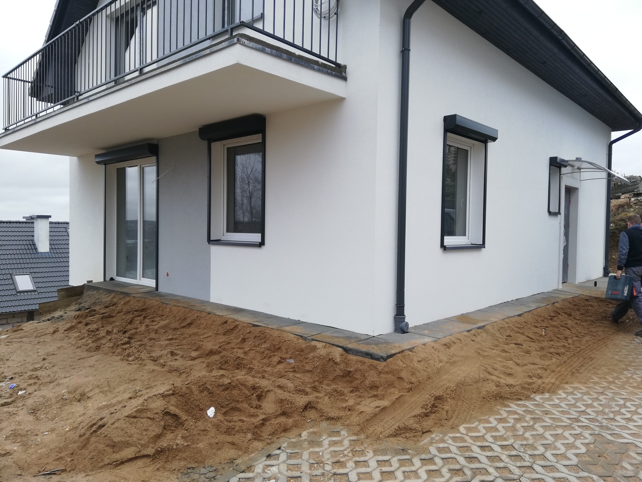 Nowo wybudowany dom z szarą elewacją, czarnymi ramami okiennymi i zewnętrznymi roletami, widok na przygotowany teren pod kostkę brukową i piasek.