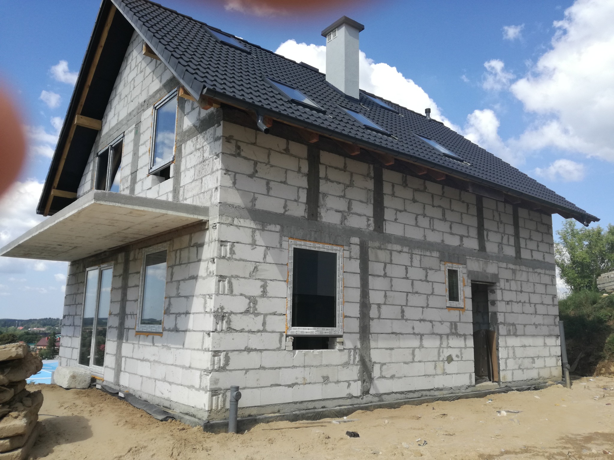 Surowy stan budynku jednorodzinnego z szarej cegły, z czarnym dachem pokrytym dachówką, widoczne okna z częściowo zamontowanymi ramami oraz komin, na tle błękitnego nieba z chmurami.