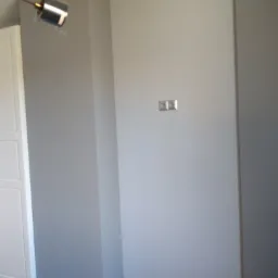 Świeżo pomalowane na szaro ściany w pokoju z drewnianą podłogą, widoczne gniazdka elektryczne i lampa sufitowa.