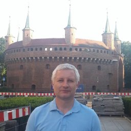 VLADISLAV RZHANSKY - Mężczyzna na tle Barbakanu w Krakowie, w tle widoczne barierki i materiały budowlane. Ujęcie z bliska, w dziennym świetle.