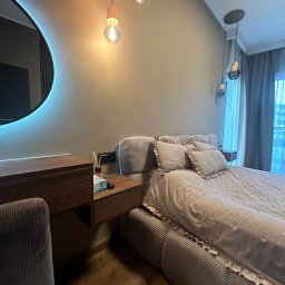 VLADISLAV RZHANSKY - Nowoczesna sypialnia w stonowanych barwach: łóżko z dekoracyjną narzutą, drewniana toaletka z okrągłym lustrem podświetlonym LED. Ciepłe światło żarówek. Parkiet na podłodze.