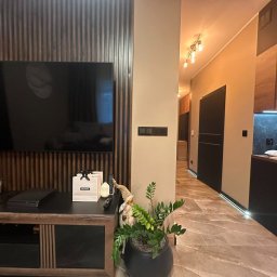 VLADISLAV RZHANSKY - Nowoczesny salon z telewizorem na ścianie z drewnianymi lamelami, marmurowa podłoga i widok na kuchnię z czarnymi meblami. Całość w ciepłych, stonowanych barwach.