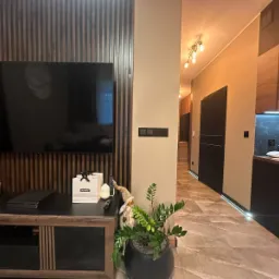 Nowoczesny salon z telewizorem na ścianie z drewnianymi lamelami, marmurowa podłoga i widok na kuchnię z czarnymi meblami. Całość w ciepłych, stonowanych barwach.