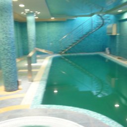 VLADISLAV RZHANSKY - Błękitny basen wewnątrz budynku z kolumnami i schodami spiralnymi, wykończony mozaiką. Oświetlenie sufitowe i refleksy w wodzie. Nietypowy design.