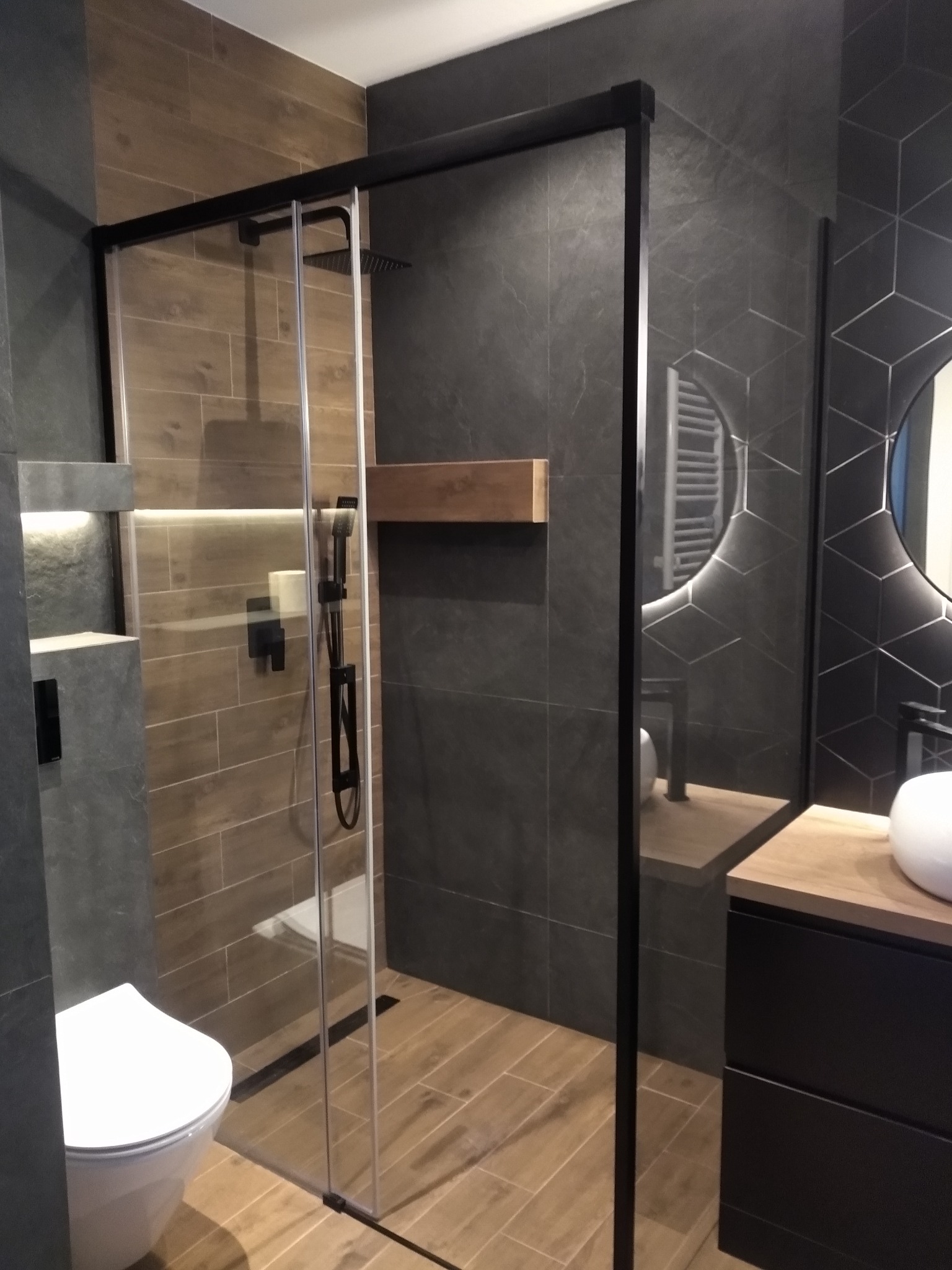 Nowoczesna łazienka z kabiną prysznicową typu walk-in z czarnymi profilami, drewnopodobnymi płytkami na ścianie i podłodze, szarą ścianą z geometrycznym wzorem oraz okrągłym lustrem...