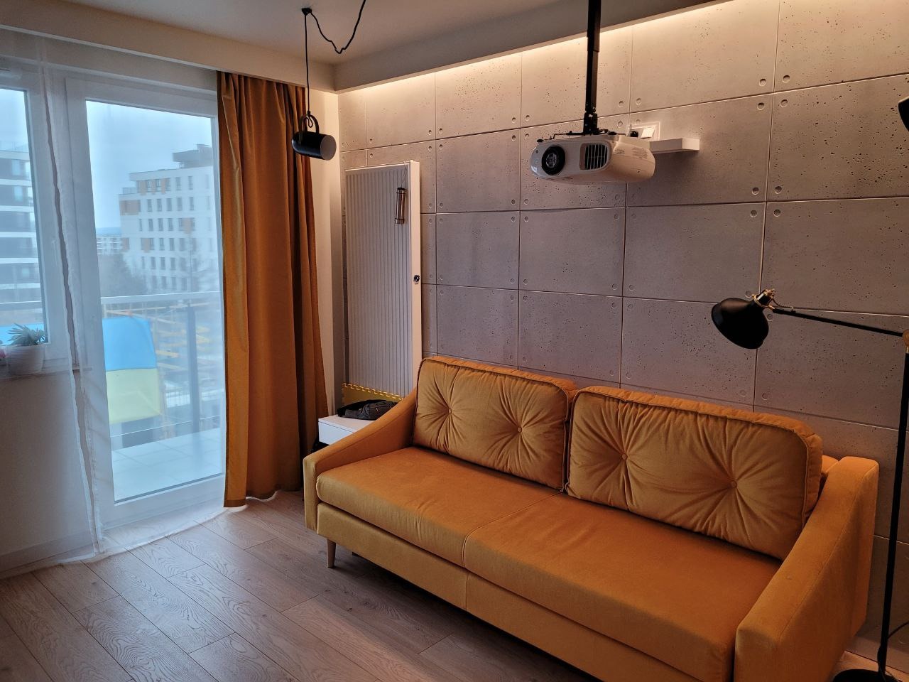 Salon z zamontowanym projektorem sufitowym, żółtą sofą na parkiecie i betonową ścianą z oświetleniem punktowym.