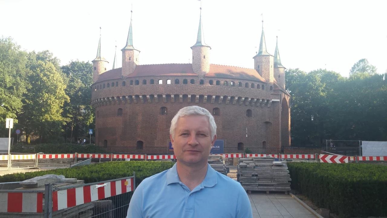 Mężczyzna na tle Barbakanu w Krakowie, w tle widoczne barierki i materiały budowlane. Ujęcie z bliska, w dziennym świetle.