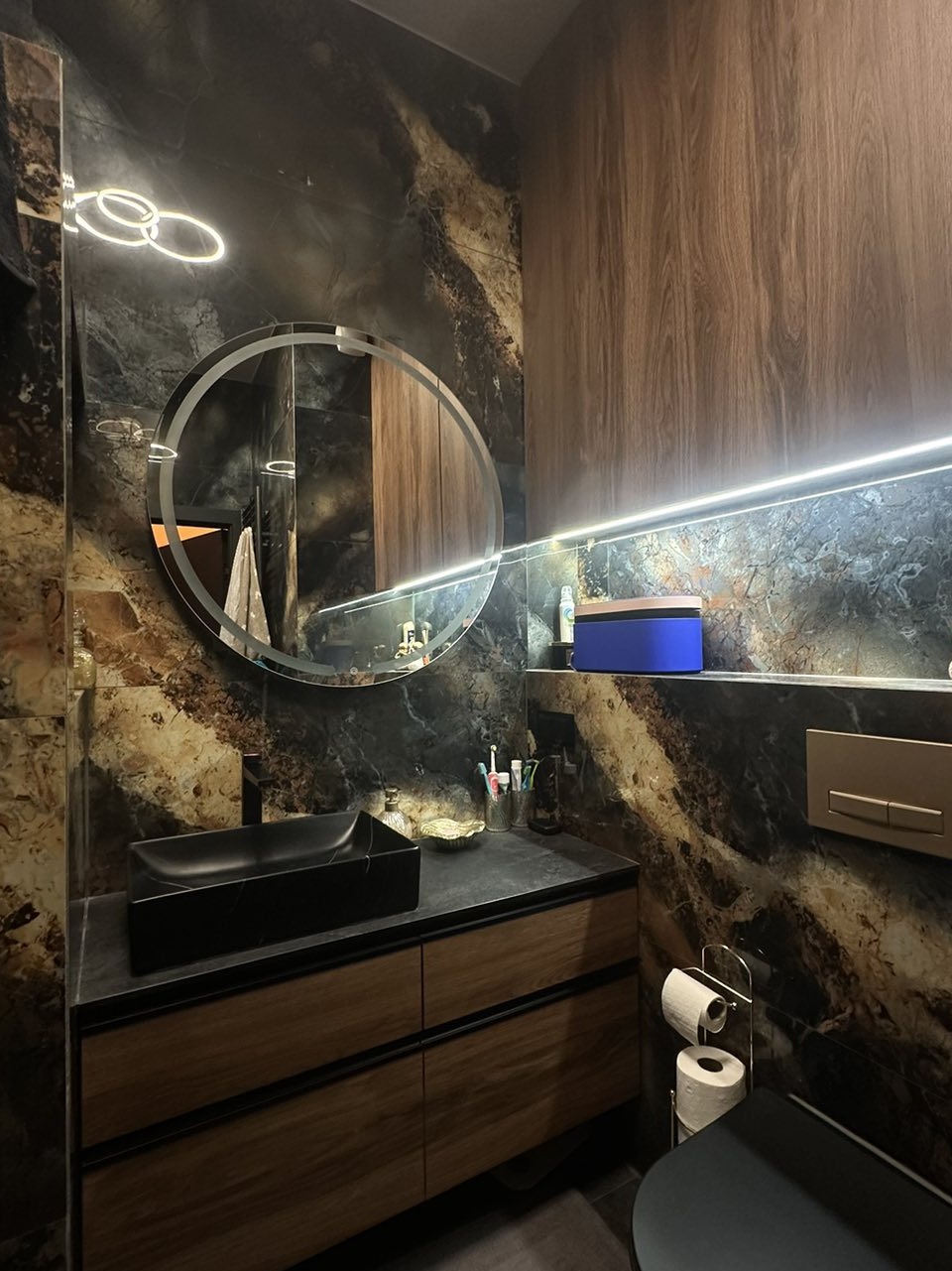 Elegancka łazienka z marmurową ścianą, okrągłym lustrem z podświetleniem LED i czarną, prostokątną umywalką na drewnianej szafce. Widoczne akcesoria łazienkowe.