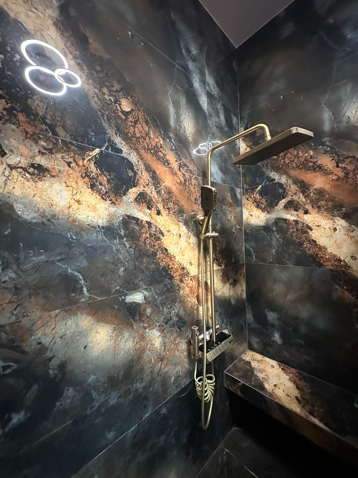 Luksusowa kabina prysznicowa z marmurowymi płytkami w odcieniach brązu i czerni, złota armatura, designerskie oświetlenie. Wykończenie z dbałością o detale.