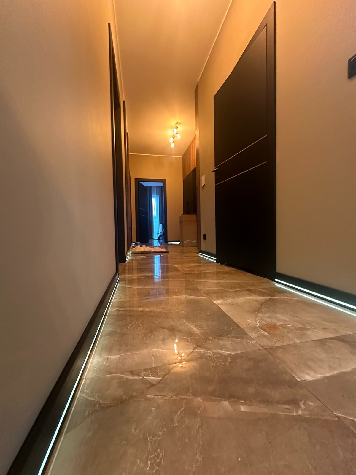 Korytarz z marmurową podłogą i oświetleniem LED przy listwach. Widoczne drzwi w kolorze ciemnego brązu z poziomymi zdobieniami oraz lampa sufitowa.