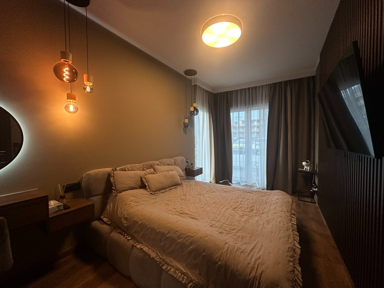 Nowoczesna sypialnia w ciepłych odcieniach beżu i brązu. Dekoracyjne lampy wiszące, telewizor na ścianie z pionowymi panelami, duże okno z zasłonami.