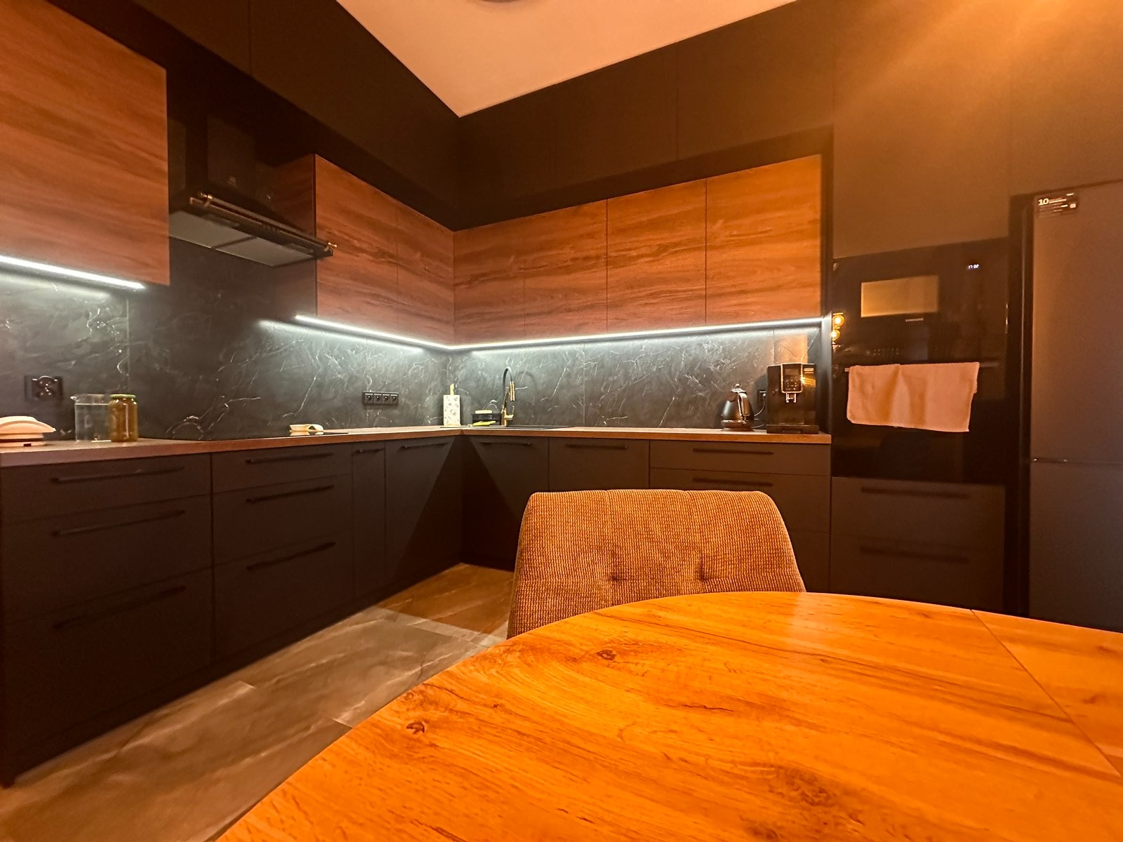 Nowoczesna kuchnia w ciemnej tonacji z drewnianymi akcentami i oświetleniem LED podszafkowym. Na pierwszym planie fragment drewnianego stołu i krzesła.