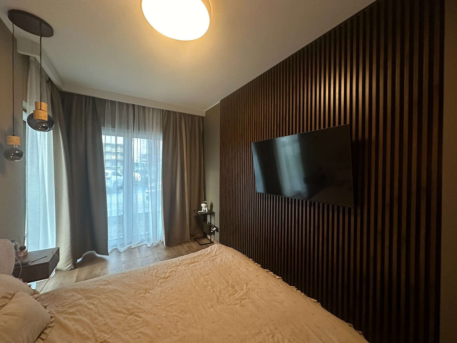 Nowoczesna sypialnia z drewnianą ścianą z lameli, na której wisi telewizor. Beżowe zasłony w oknie i łóżko w odcieniach beżu. Designerskie lampy wiszące.