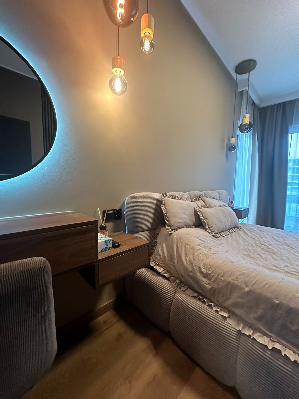 Nowoczesna sypialnia w stonowanych barwach: łóżko z dekoracyjną narzutą, drewniana toaletka z okrągłym lustrem podświetlonym LED. Ciepłe światło żarówek. Parkiet na podłodze.