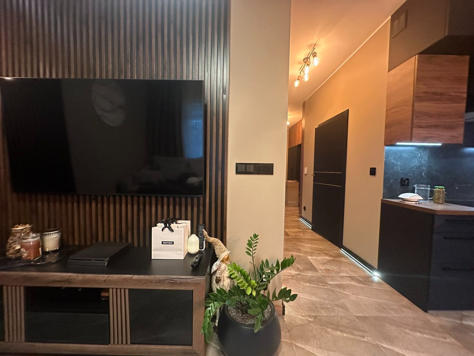 Nowoczesny salon z telewizorem na ścianie z drewnianymi lamelami, marmurowa podłoga i widok na kuchnię z czarnymi meblami. Całość w ciepłych, stonowanych barwach.