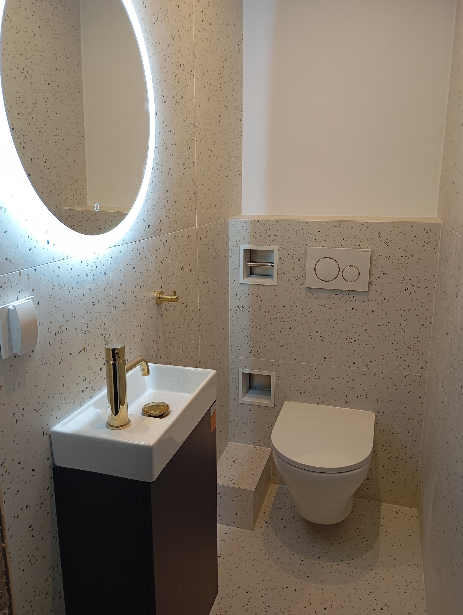 Nowoczesna łazienka z umywalką na szafce, toaletą podwieszaną i okrągłym lustrem z podświetleniem LED. Płytki w stylu lastryko na ścianach i podłodze.