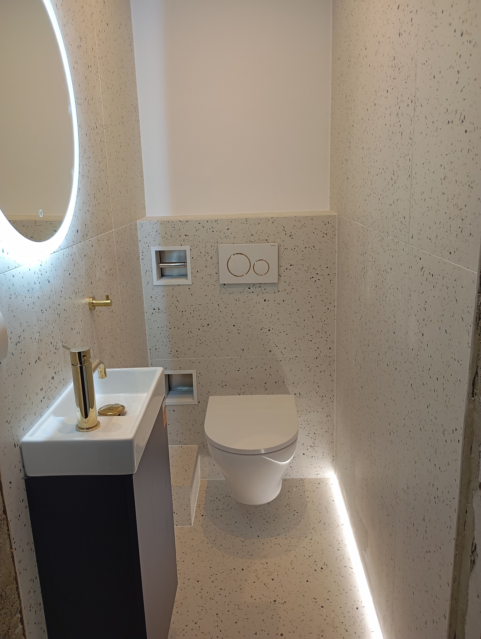 Nowoczesna łazienka z umywalką nablatową, złotą armaturą, podświetlanym lustrem i toaletą podwieszaną. Płytki w stylu lastryko na ścianach i podłodze, z ledowym oświetleniem podłogowym.