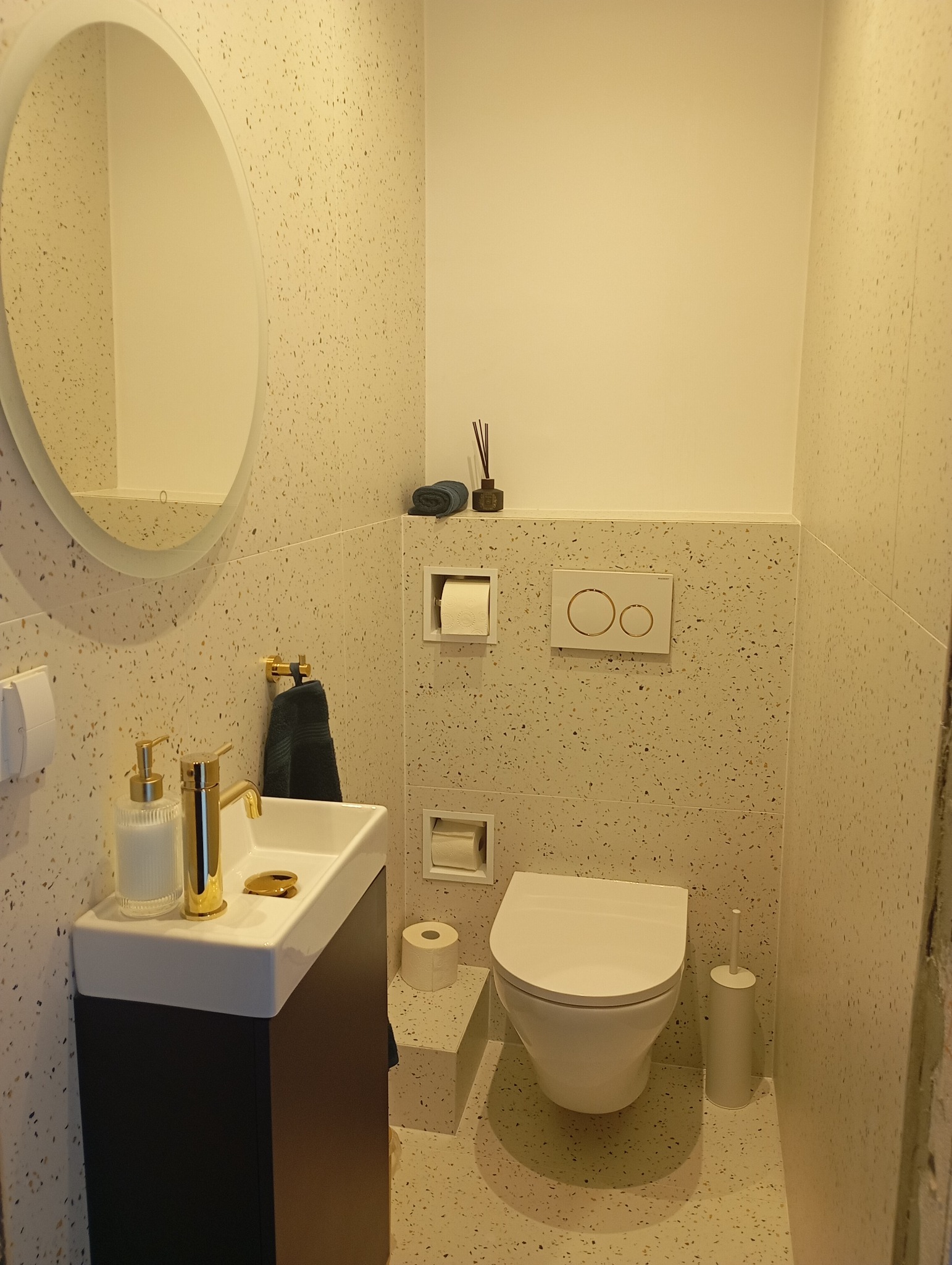 Wykończona łazienka w beżowych tonacjach z umywalką na szafce, okrągłym lustrem i toaletą. Na ścianach i podłodze wzór lastryko. Złote akcenty armatury.