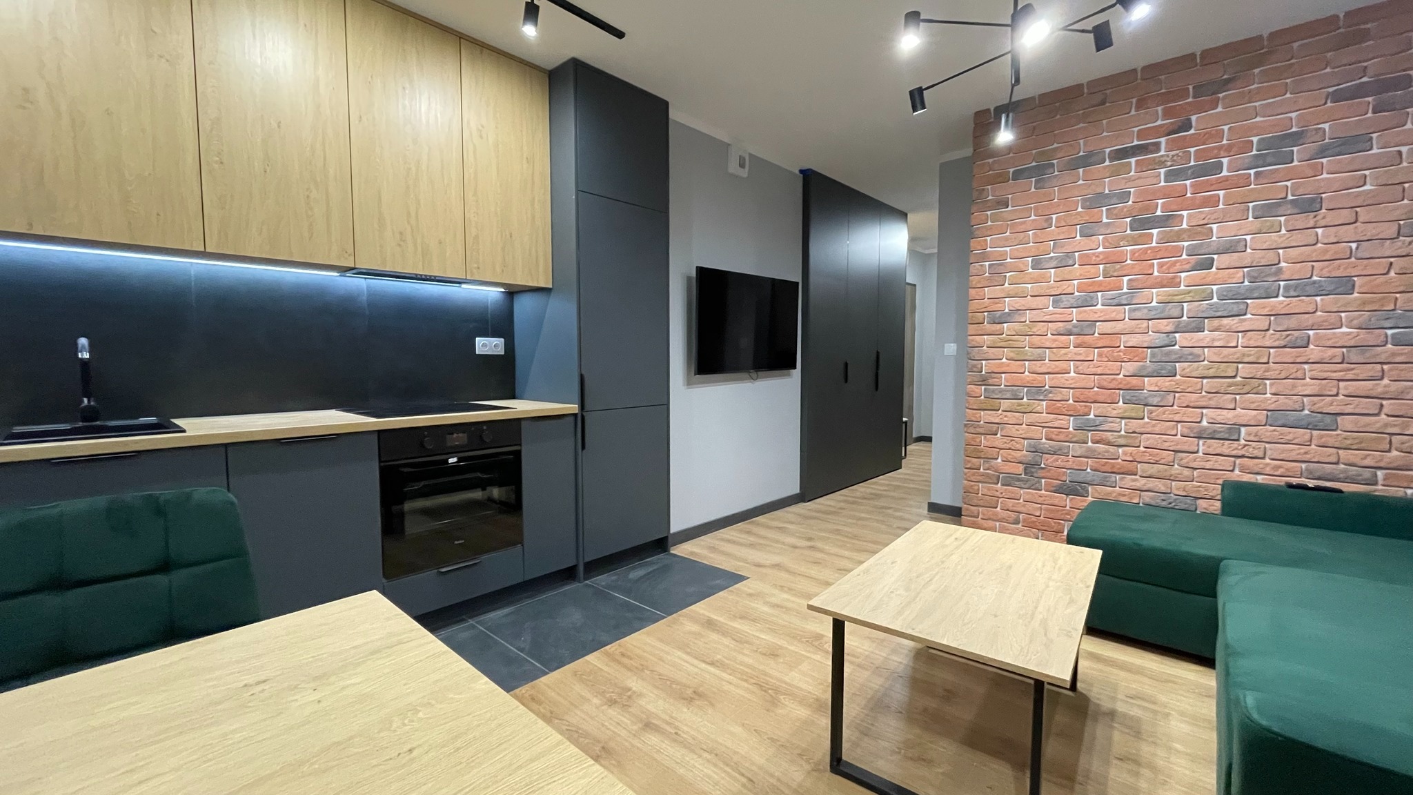 Nowoczesne wnętrze: szara zabudowa kuchenna z drewnianymi szafkami, ceglana ściana, drewniana podłoga i stół. Minimalistyczny design z oświetleniem LED.