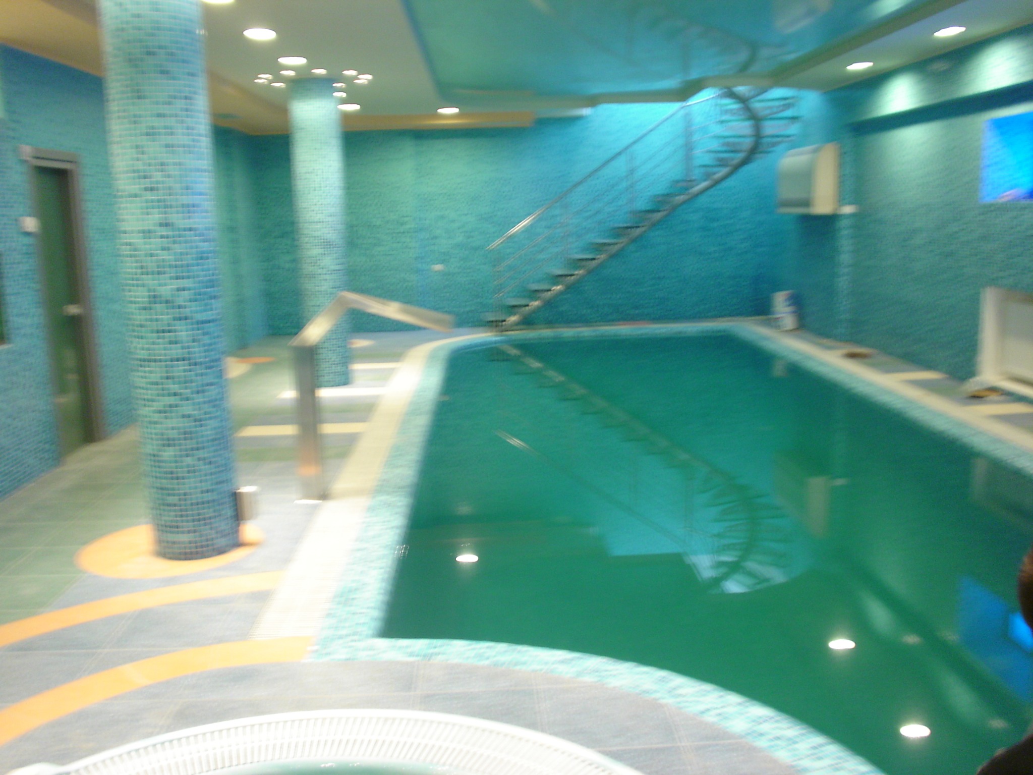 Błękitny basen wewnątrz budynku z kolumnami i schodami spiralnymi, wykończony mozaiką. Oświetlenie sufitowe i refleksy w wodzie. Nietypowy design.