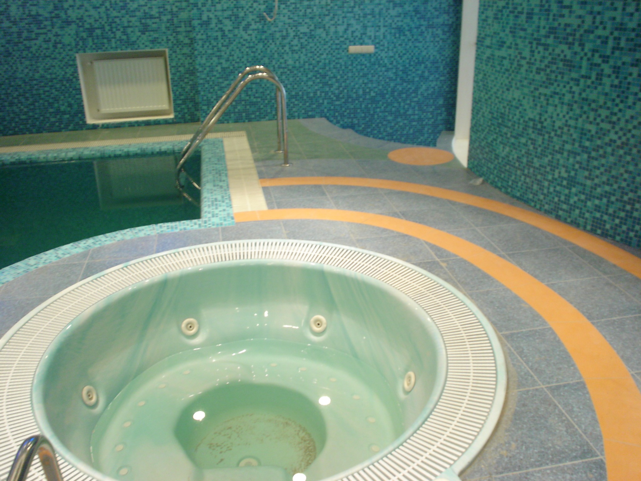 Błękitna mozaika i jacuzzi w łazience. Wykończenie wnętrza z akcentem na relaks i wodne atrakcje. Płytki podłogowe z pomarańczowymi akcentami.