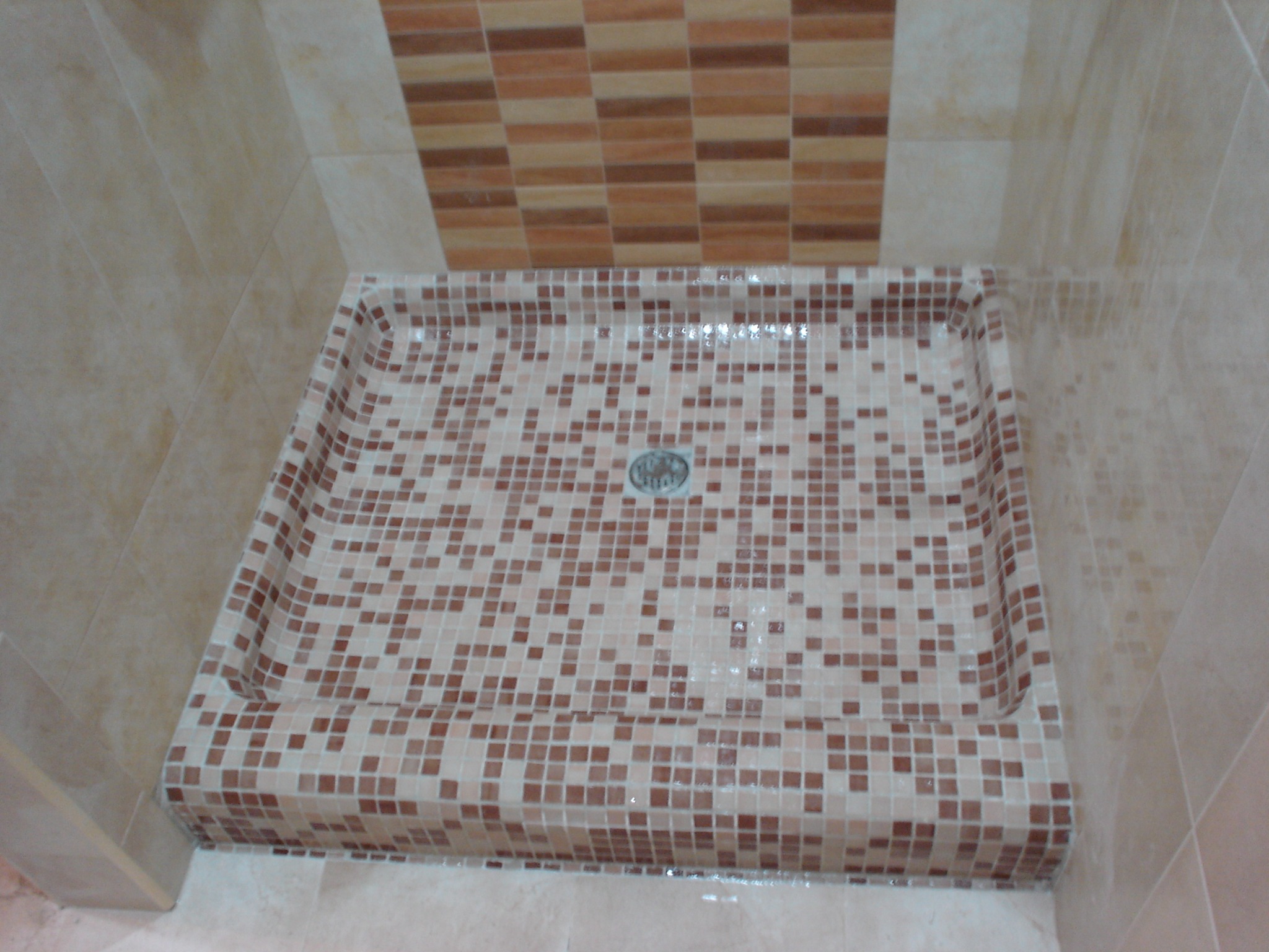 Brodzik mozaikowy w odcieniach beżu i brązu, z odpływem centralnym, otoczony jasnymi płytkami. U góry dekoracyjny pas płytek w podobnej kolorystyce.