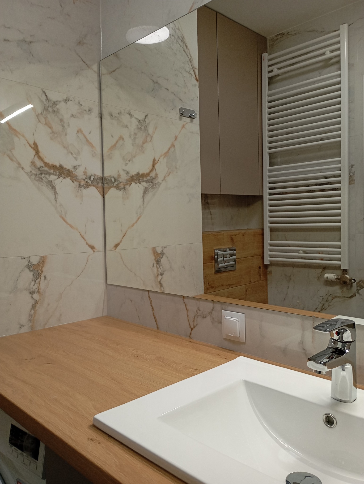 Wykończona łazienka z marmurowymi płytkami, drewnianym blatem i białą umywalką. Widoczny grzejnik drabinkowy i szafka w neutralnym odcieniu. Nowoczesny design.