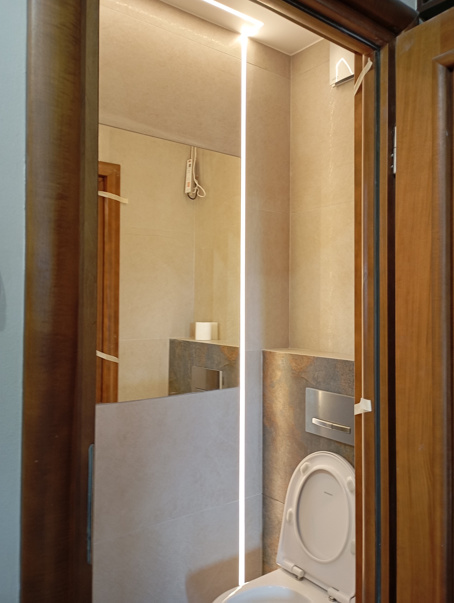 Wykończona łazienka z podświetlanym lustrem i toaletą w neutralnych barwach, widoczna przez drewniane futryny drzwi. Minimalistyczny design z elementami dekoracyjnymi.