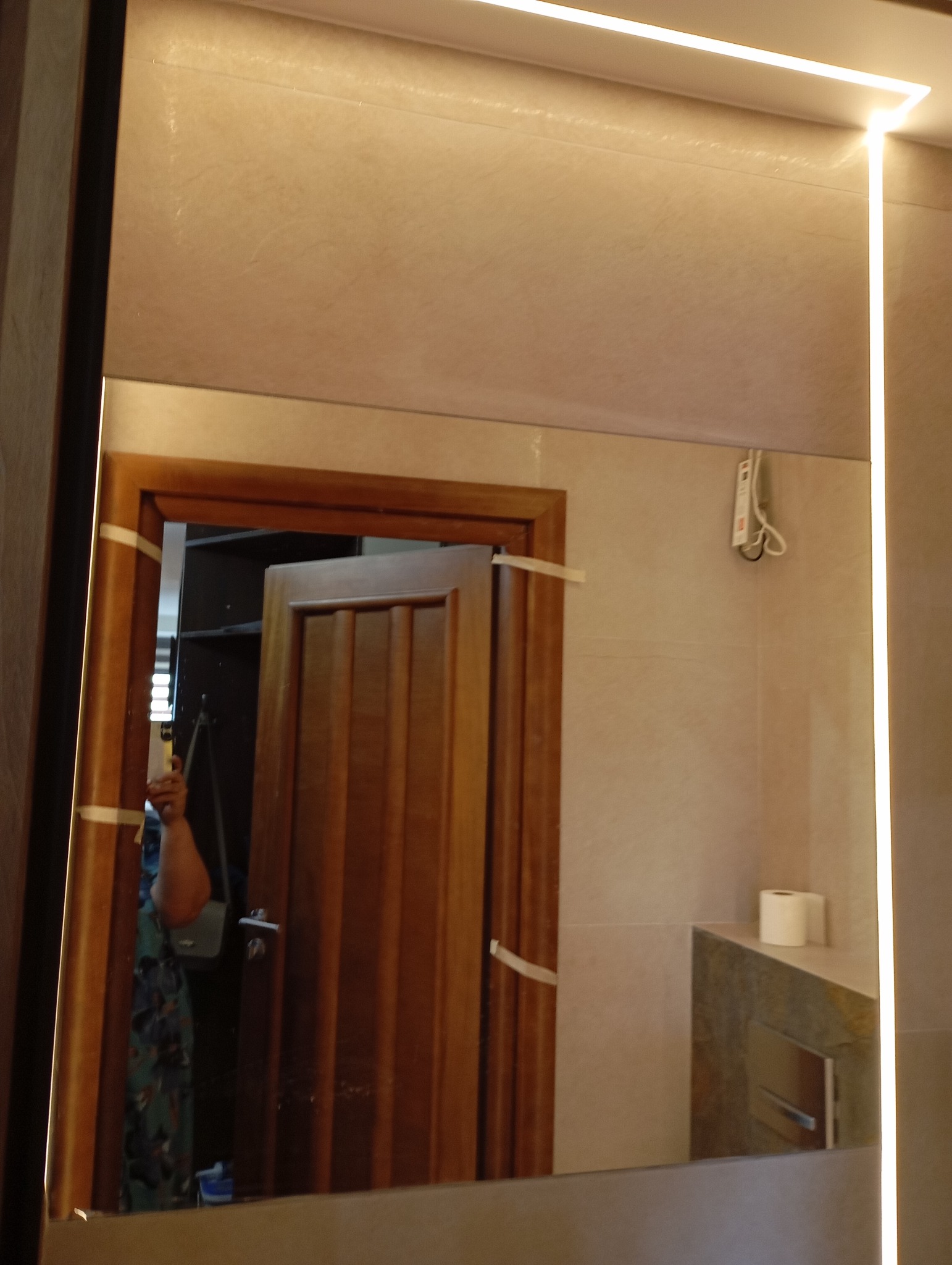 Nowoczesne lustro łazienkowe z podświetleniem LED, odbijające fragment drzwi i wnętrze. Widoczne płytki w neutralnych barwach i elementy wyposażenia łazienki.